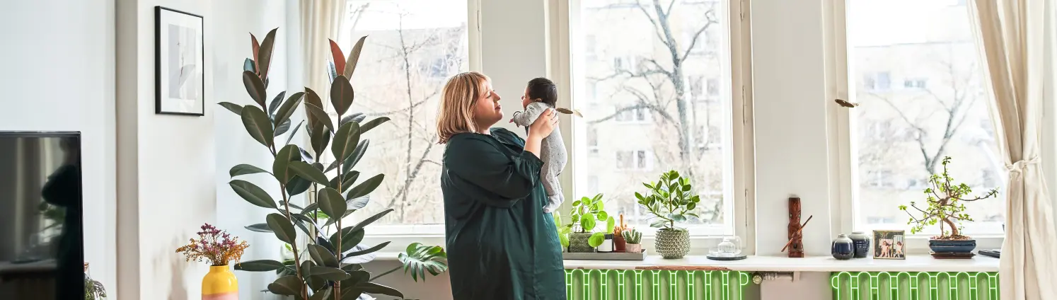 Eine Mutter schaut ihr Kind in den Armen an. Sie steht im Wohnzimmer.