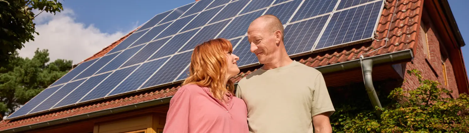 Ein lächelndes Pärchen steht vor ihrem Haus und das Dach ist mit einer PV-Anlage ausgestattet.