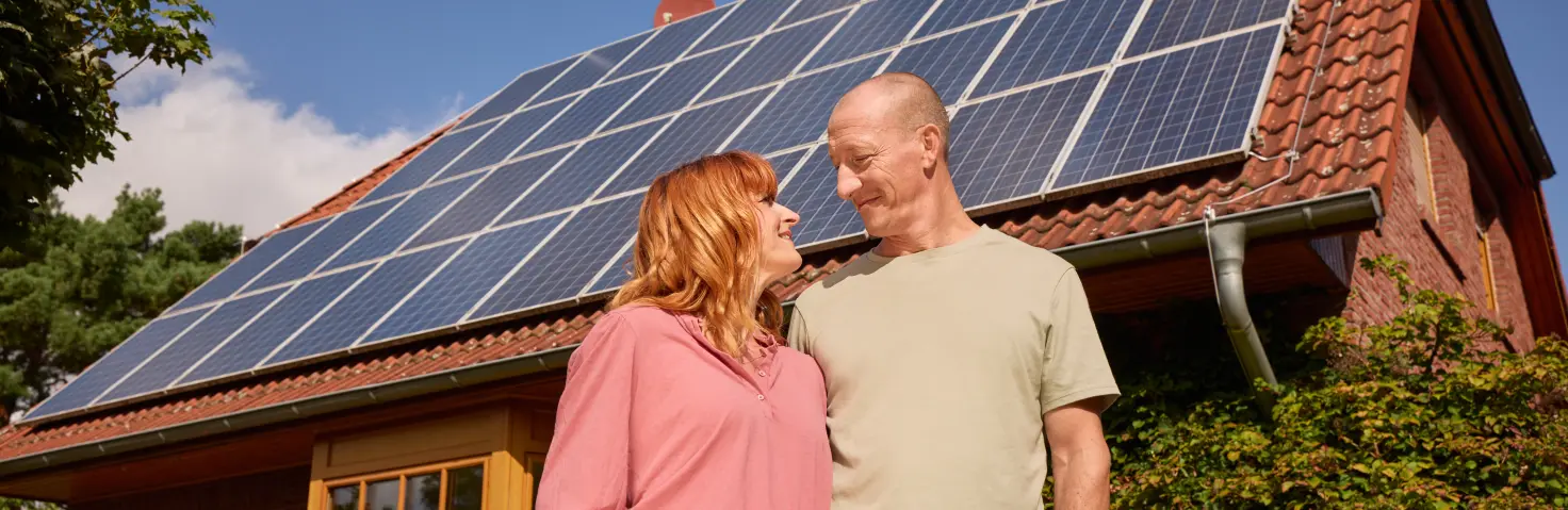 Ein lächelndes Pärchen steht vor ihrem Haus und das Dach ist mit einer PV-Anlage ausgestattet.