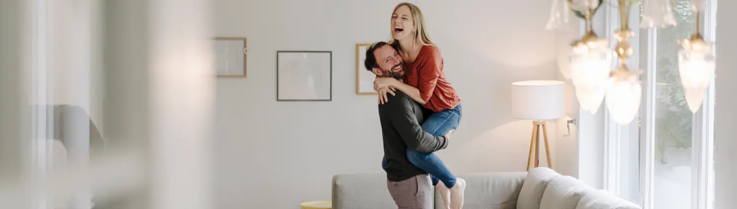 Ein Pärchen freut sich in einer hellen Wohnung vor einem Sofa, der Mann hebt die Frau hoch