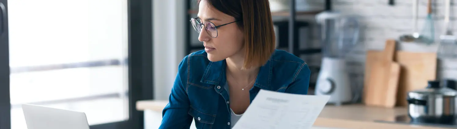 Frau mit Brille die mit dem Computer arbeitet, während sie einige Rechnungen und Dokumente in der Küche zu Hause liest