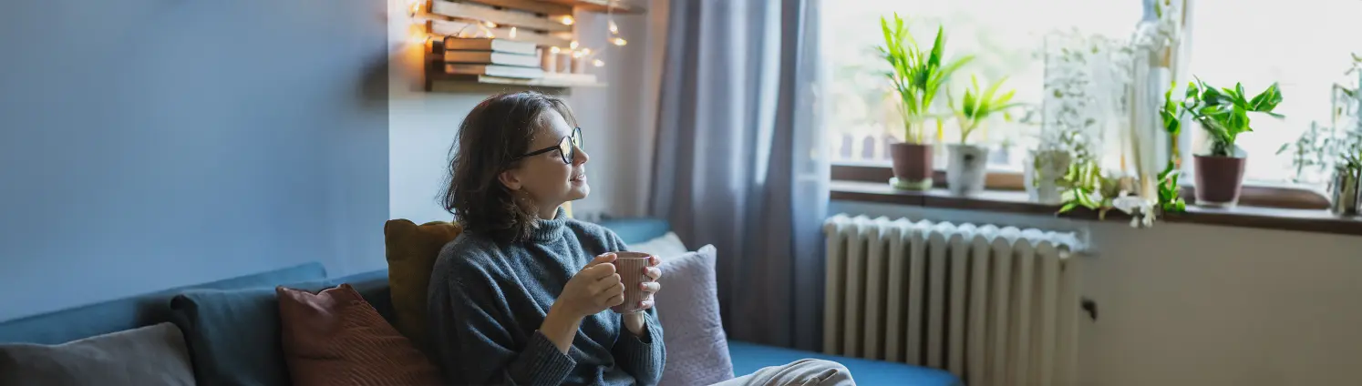 Eine Frau mit Tasse in der Hand sitzt auf ihre Sofa und lächelt