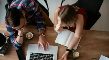 Eine Frau und ein Mann sitzen an einem Tisch und beugen sich über einen Laptop und Papiere