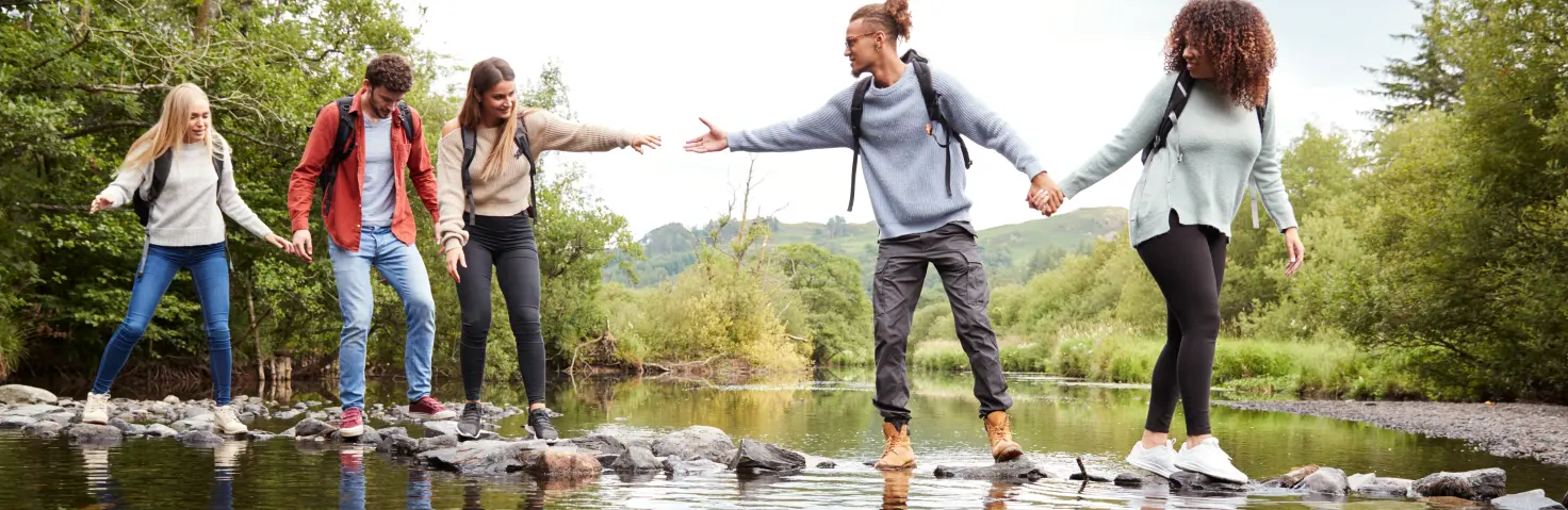 Junge Menschen helfen sich bei einer Wanderung gegenseitig über einen Flusslauf.