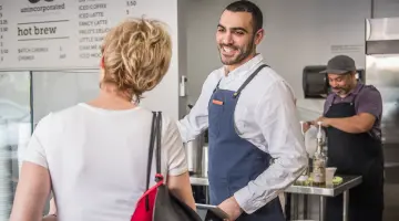 Gewerbestrom: Sympathischer Barista in einem stylischen Coffee Shop bedient lächelnd eine Kundin.