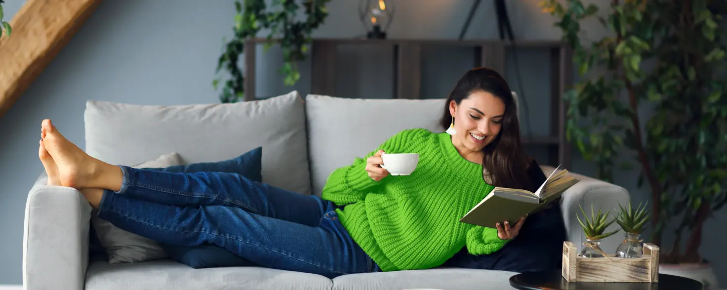 Eine Frau liegt mit einer Tasse Tee auf dem Sofa und liest ein Buch