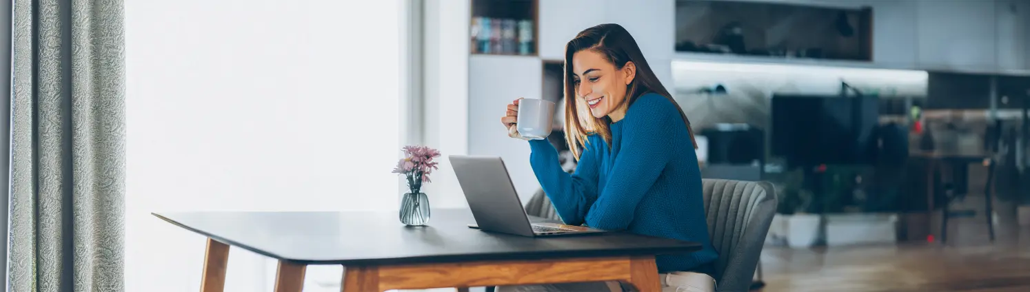 Eine Frau mit Tasse in der Hand schaut lächelnd auf ihren Laptop