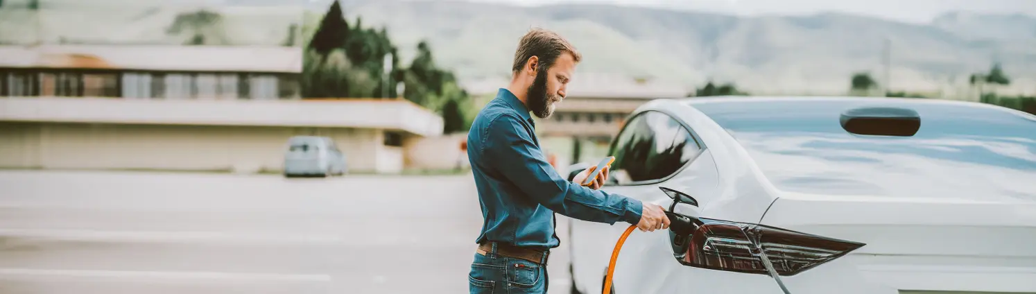 Ein Mann lädt ein Elektroauto auf und schaut dabei auf sein Handy