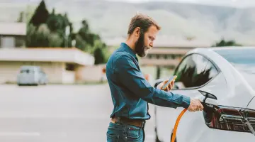 Ein Mann lädt ein Elektroauto auf und schaut dabei auf sein Handy