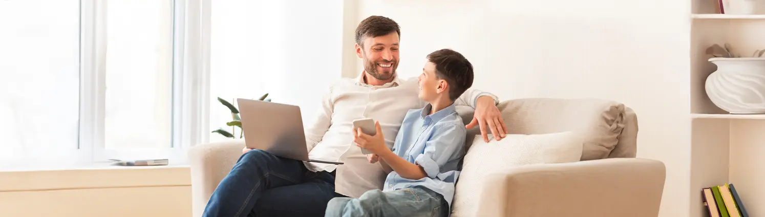 Vater und Sohn sitzen lachend zusammen auf einem Sofa mit Laptop auf dem Schoß