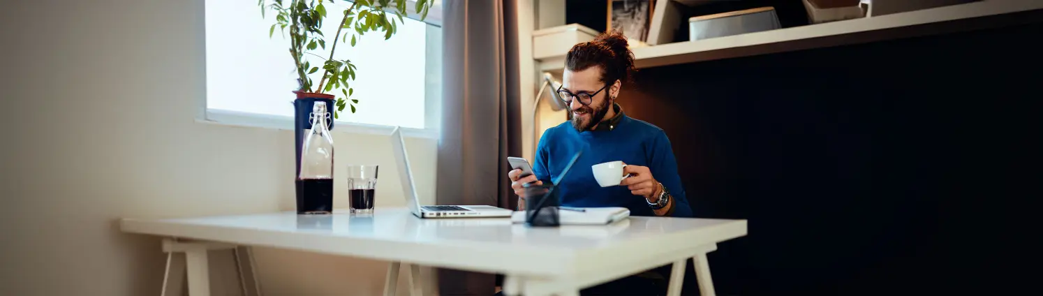 Ein Mann sitzt mit seinem Handy und einer Tasse in der Hand vor seinem Laptop