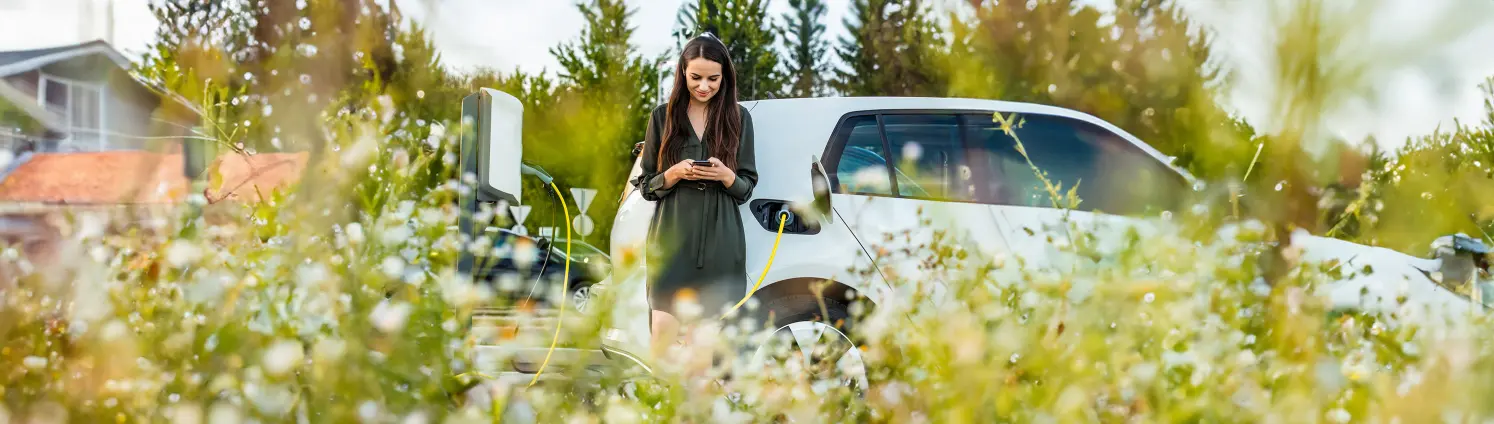 Junge Frau im Kleid lädt ihr E-Auto und schaut beim Warten auf ihr Handy.