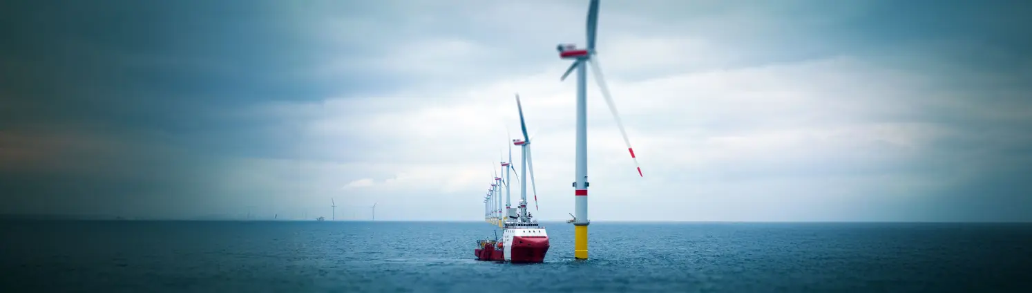 Mehrere Windräder stehen in einer Linie auf dem Meer, daneben fährt ein Schiff