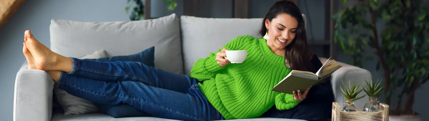 Eine Frau liegt auf ihrem Sofa, trinkt Tee und ließt ein Buch