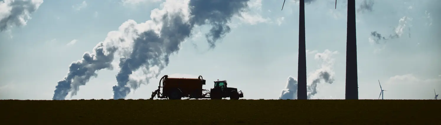 Ein Feld an dessen Horizont Windkrafträder zu sehen sind, ein Traktor fährt über das Feld und Rauchschwaden sind zu sehen