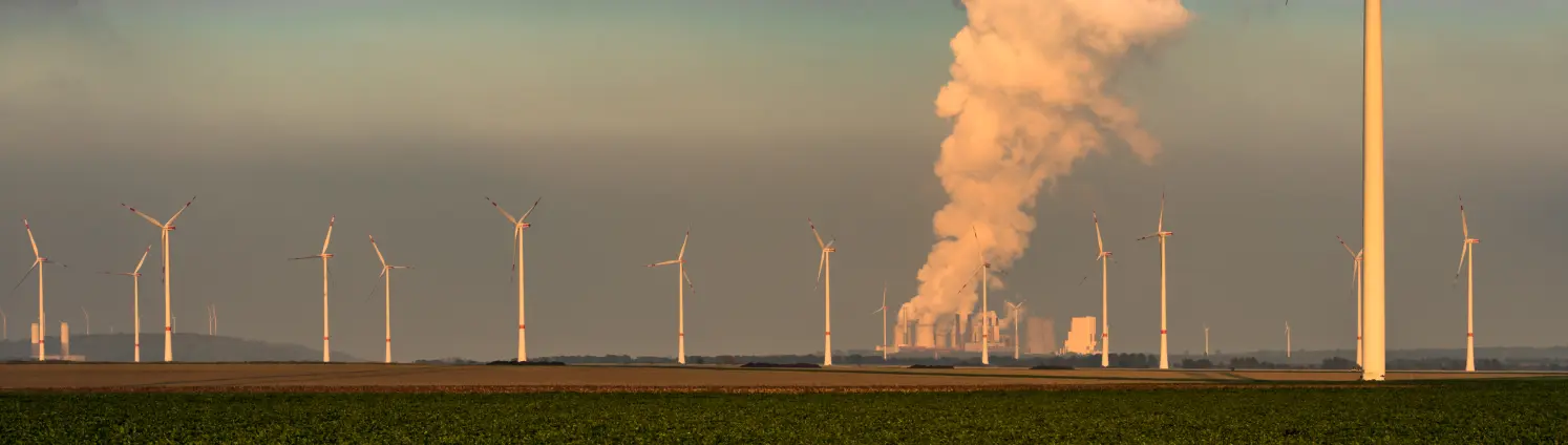 Windräder auf einem Feld. Im Hintergrund steigt Abgas aus dem Schornstein eines Kraftwerks.