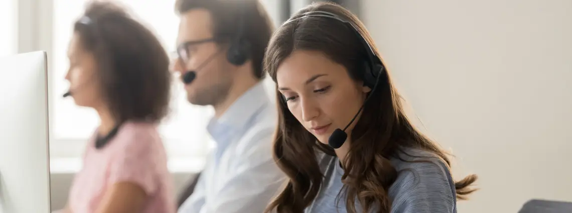 Blick auf ein Callcenter, eine Frau mit Headset ist im Vordergrund, sie liest einen Zettel