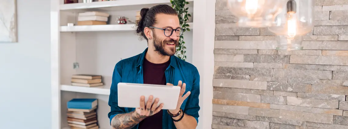 Ein Mann mit Brille und Bart steht vor einem Bücherregal, hält ein Tablet in der Handy und schaut lachend zur Seite