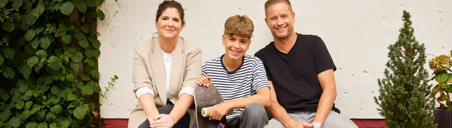 Mutter, Sohn mit Skateboard und Vater sitzen auf einer Bank vor ihrem Einfamilienhaus.