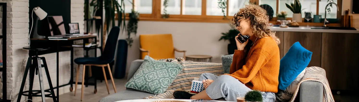 Frau sitzt mit orangen Pullover auf der Couch und Telefoniert. Dabei lächelt sie.