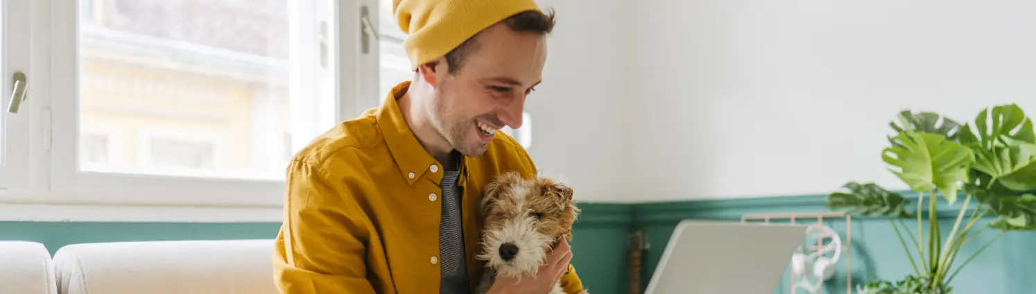 Junger Mann mit Beanie sitzt mit seinem Foxterrier auf dem Sofa und blickt auf seinen Laptop.